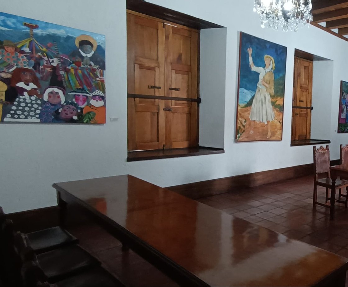 Salón con una larga mesa de madera en primer plano, ventanas de contraventanas de madera y grandes pinturas costumbristas colgadas en la pared.
