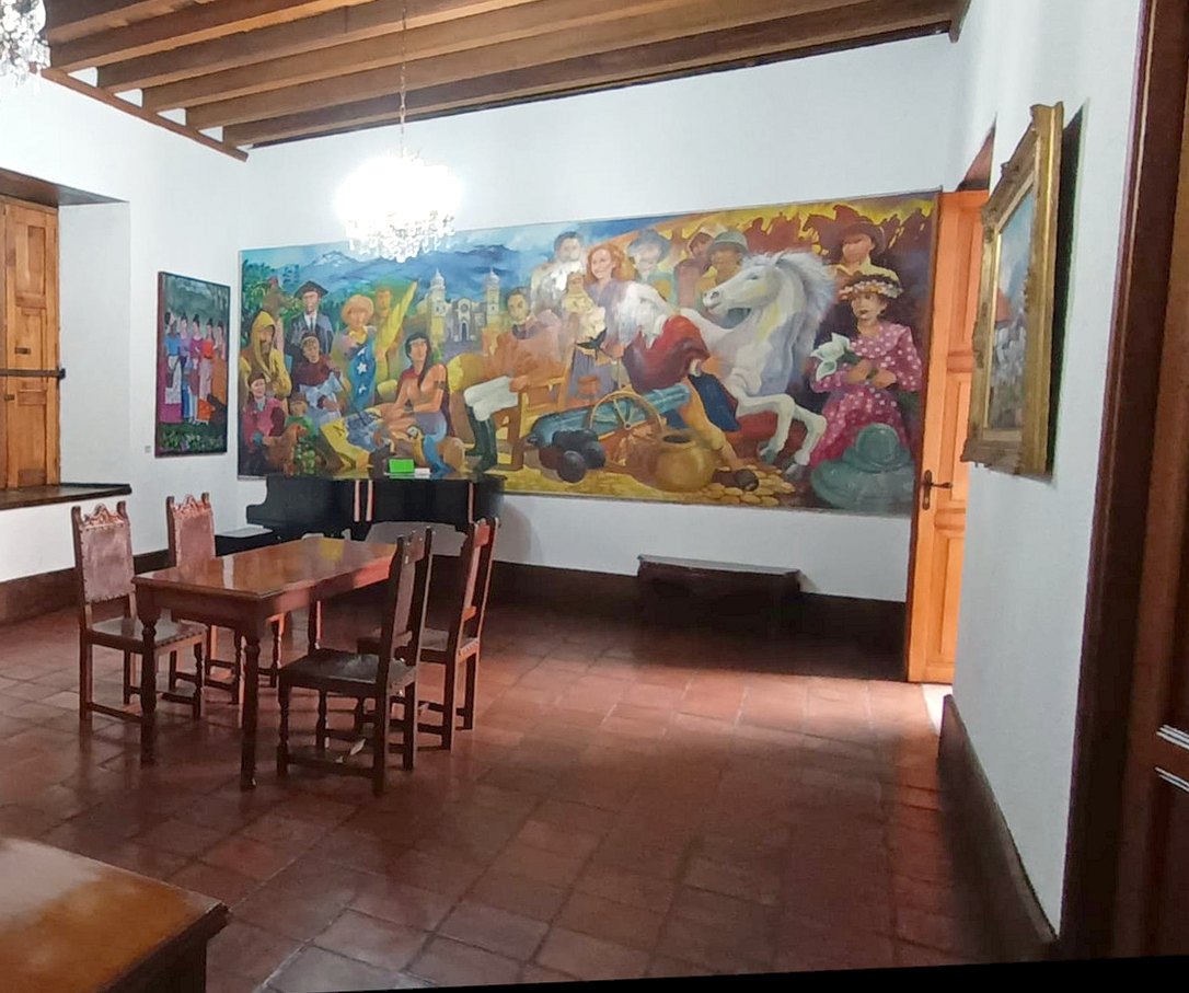 Piano de cola negro junto a un mural colorido de escena popular, con una mesa y sillas de madera sobre el piso de baldosas de barro.